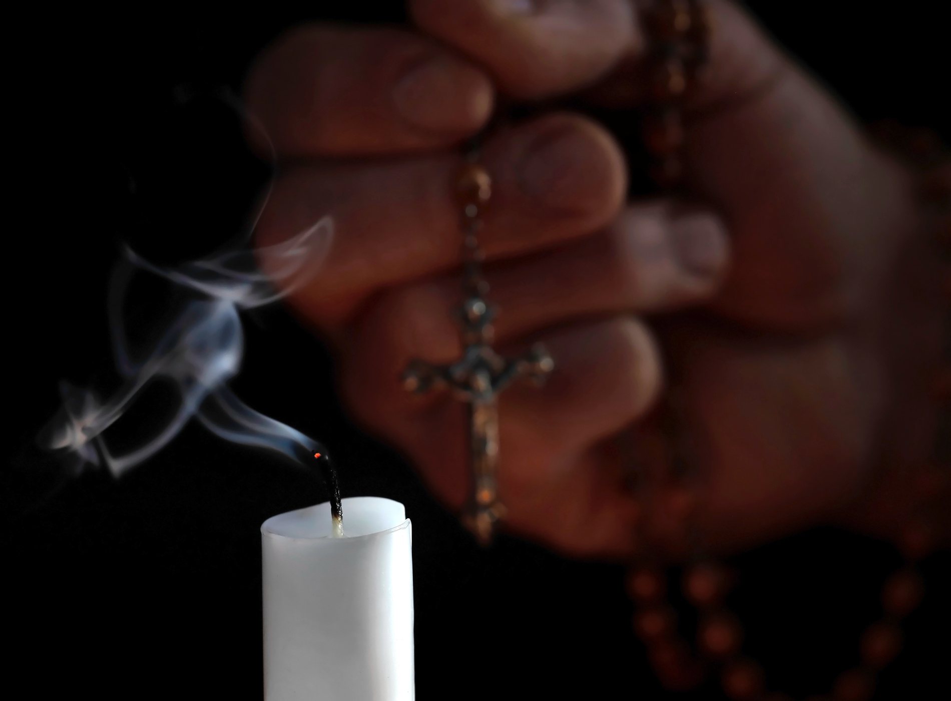 Blown out white candle with smoke in front of male hand with rosary on black background, the extinguished candle symbolizes the last prayer for a deceased person and belief in life after death.