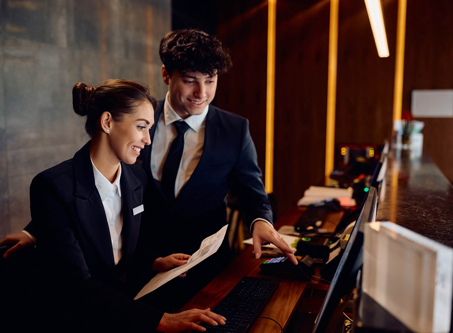 Happy hotel managers through reservation list on desktop PC at reception desk.