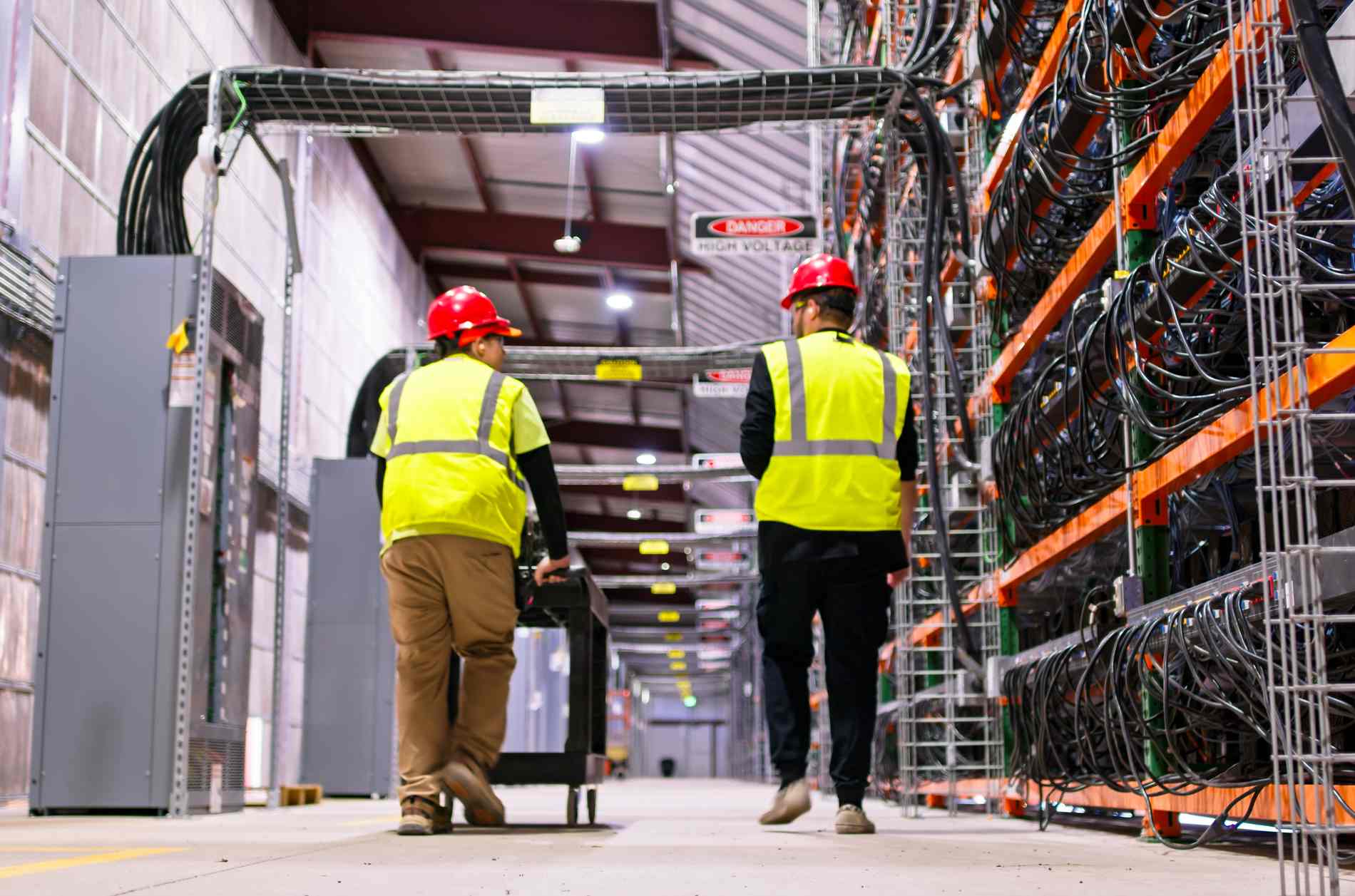 Two technicians wearing PPE walking past racks of equipment in a data center for cryptocurrency mining, cloud services and AI computing in a large, temperature controlled warehouse in a remote location in  Stutsman County, North Dakota. One of them is pushing a cart.