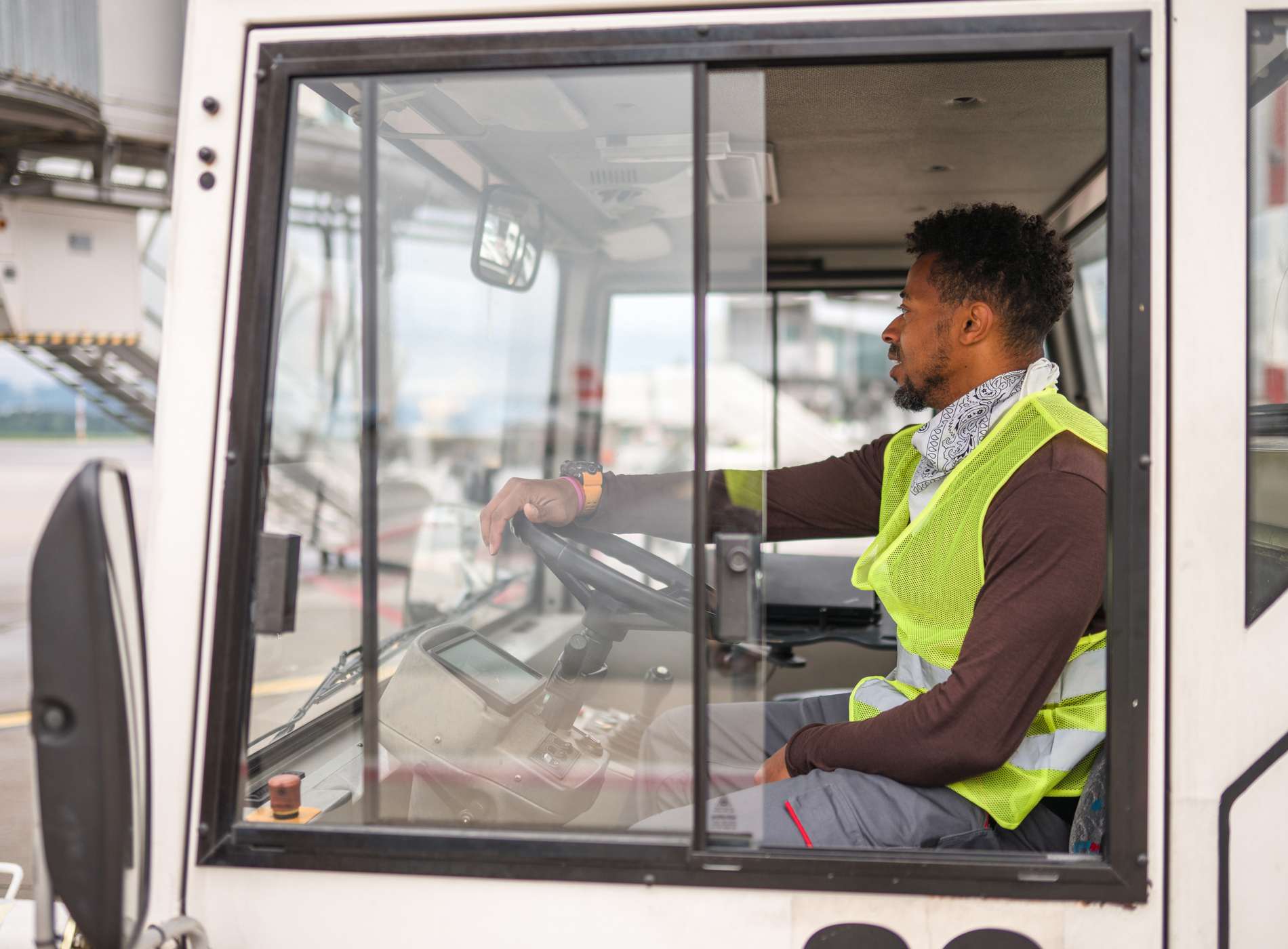Side view of a mixed race male worker driving a truck at the airport. He is placing a staircase close to a plane. He is wearing a reflective uniform.