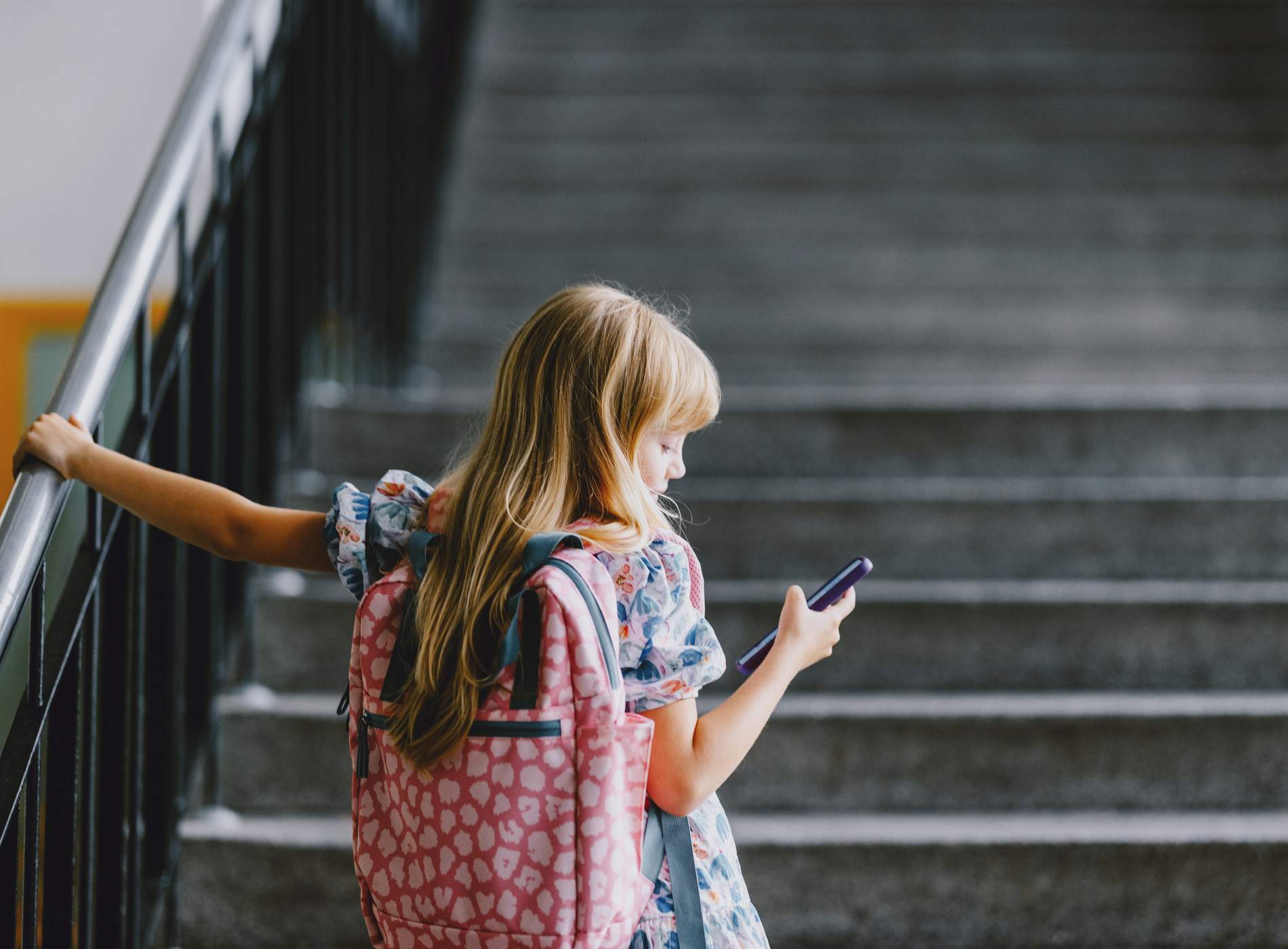 A young girl with a backpack holds her phone while standing on a school staircase. Back to school concept.