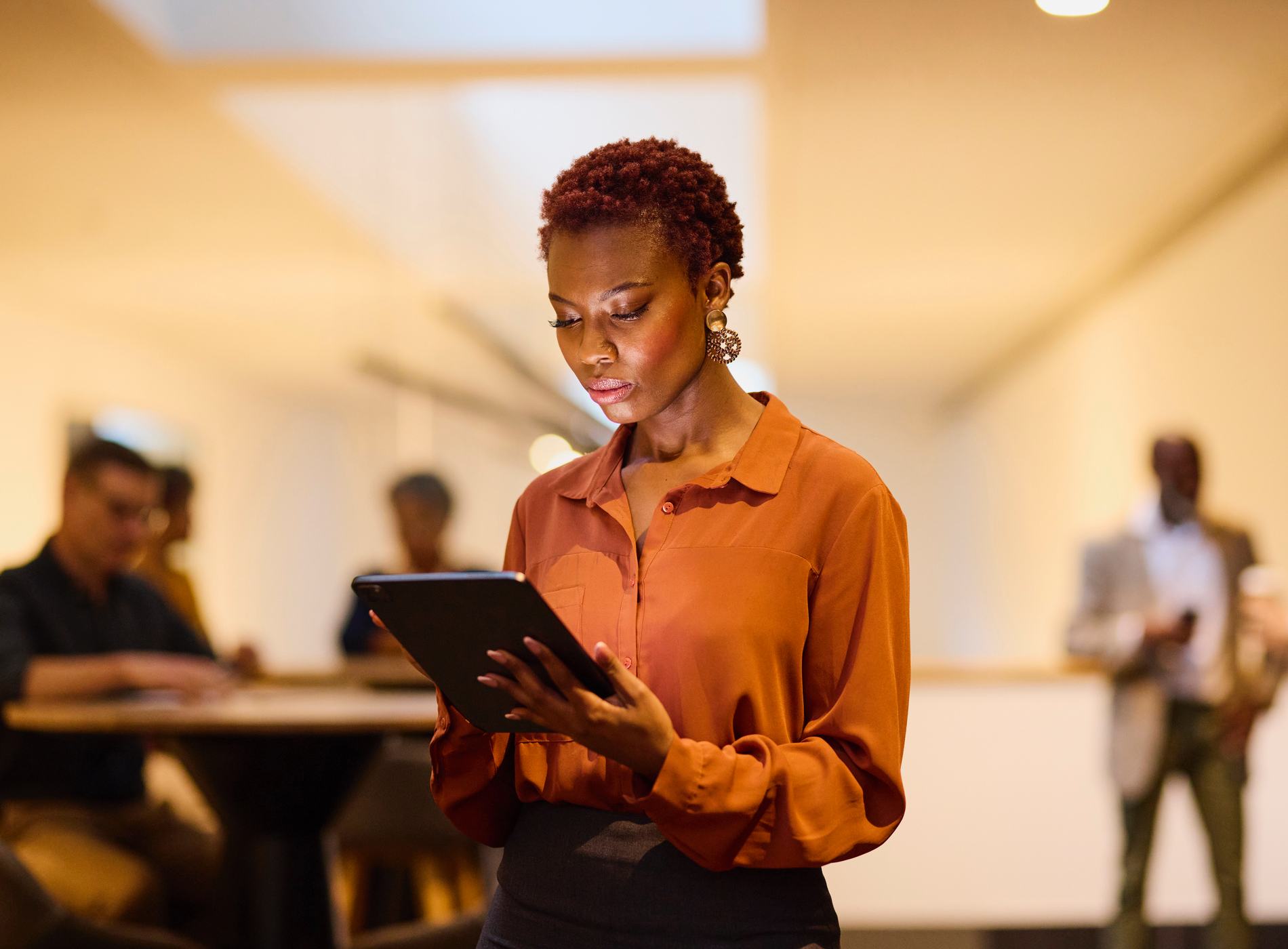 A stylish businesswoman intently reviews information on her tablet. Soft focus colleagues in the background suggest a dynamic office environment.