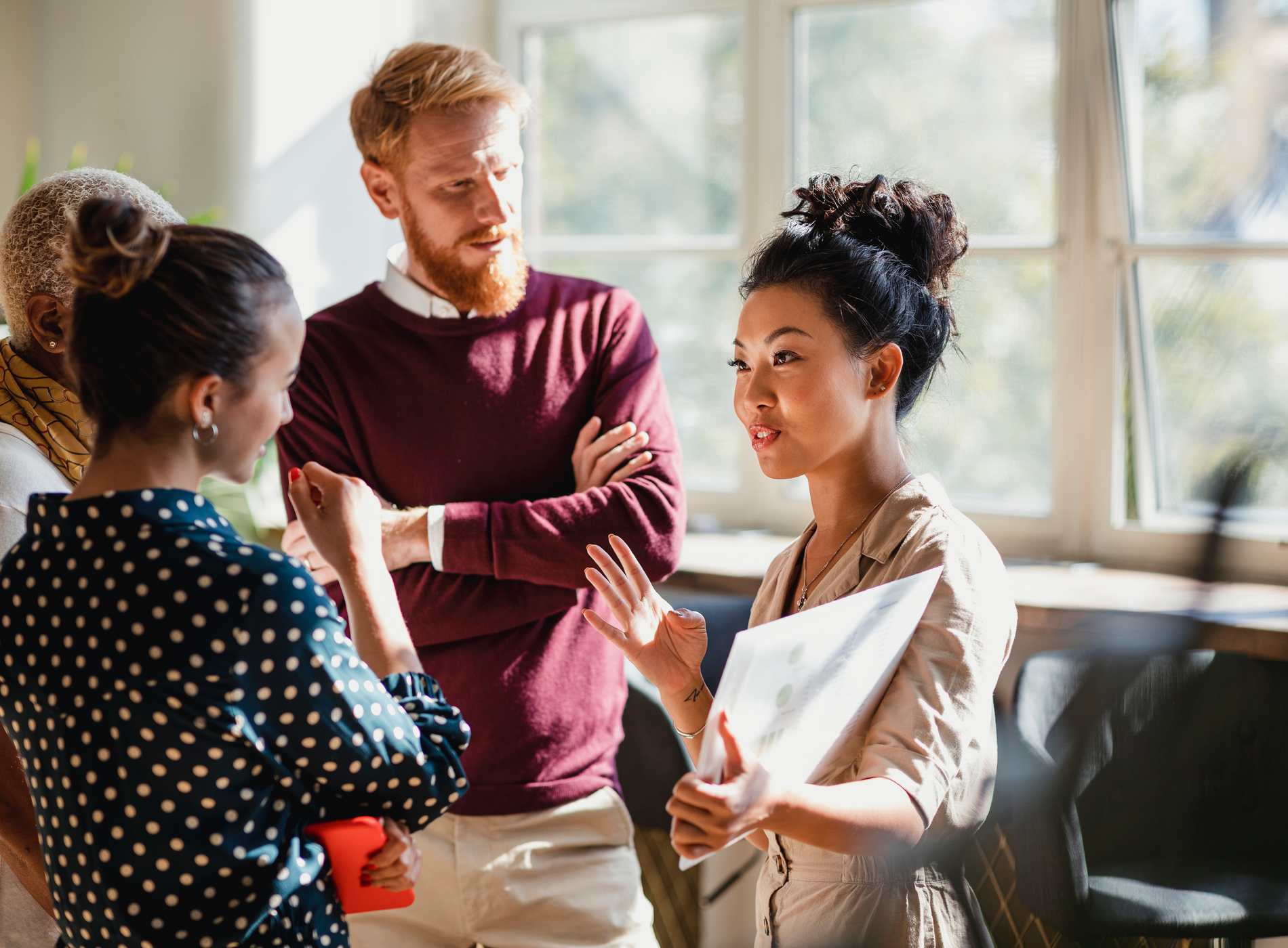 Colleagues standing in a small group discussing something. One of the women is holding documents and gesturing with her hands as the others watch and listen.