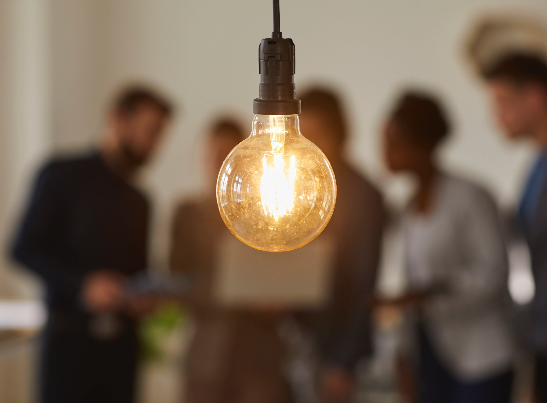 Glowing light bulb and business people over blurred background. Hanging light bulb on foreground, team of business people discussing ideas and brainstorming. Bright idea and creativity concept