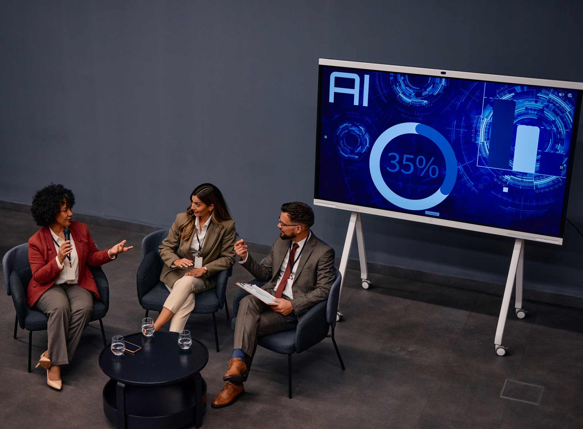 Three businesspeople are sitting in armchairs and discussing artificial intelligence during a summit, with a large screen displaying AI data in the background