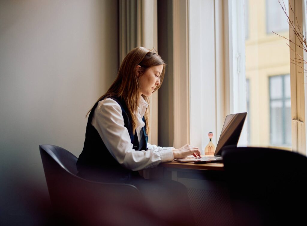 Side view of a young professional woman concentrating on her work at a wooden desk with a laptop, natural light streaming through a large window in a modern office setting.