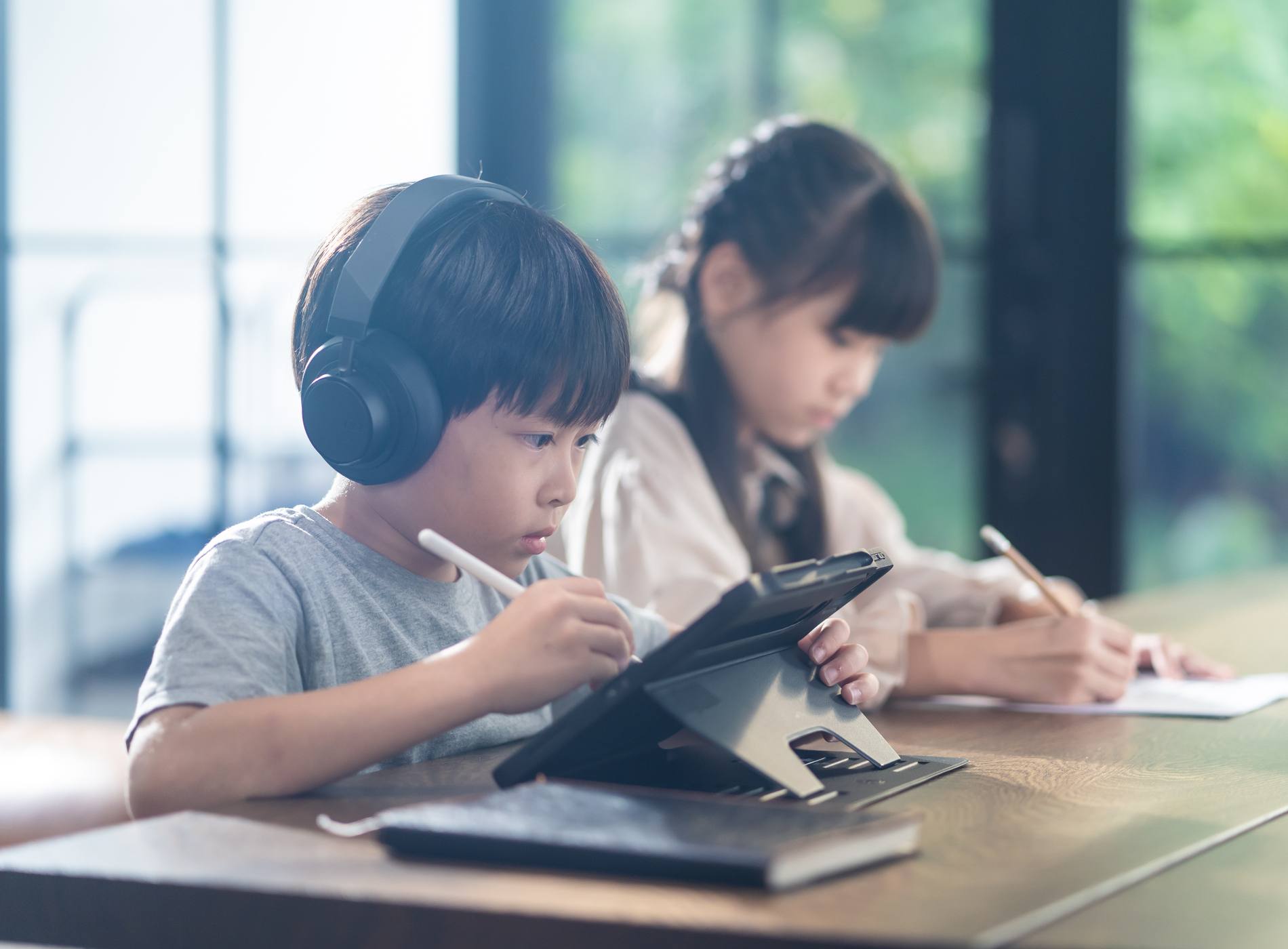 Asian little young children self learning at home. The boy using tablet and headphones for E-learning at home. The girl doing homework writing note on the paper sheet. Education and technology concept