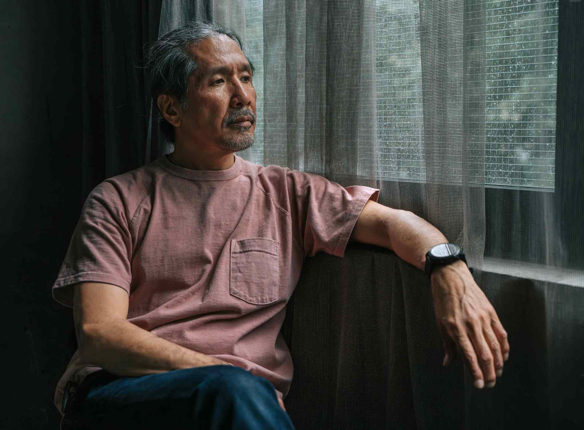 A thoughtful mature Japanese man sits quietly by a window on a rainy day, gazing outside. Dressed in a pink T-shirt and jeans, with natural light softly filtering through sheer curtains, the scene conveys introspection, solitude, and emotional depth. Ideal for themes of reflection, aging, and peaceful moments.