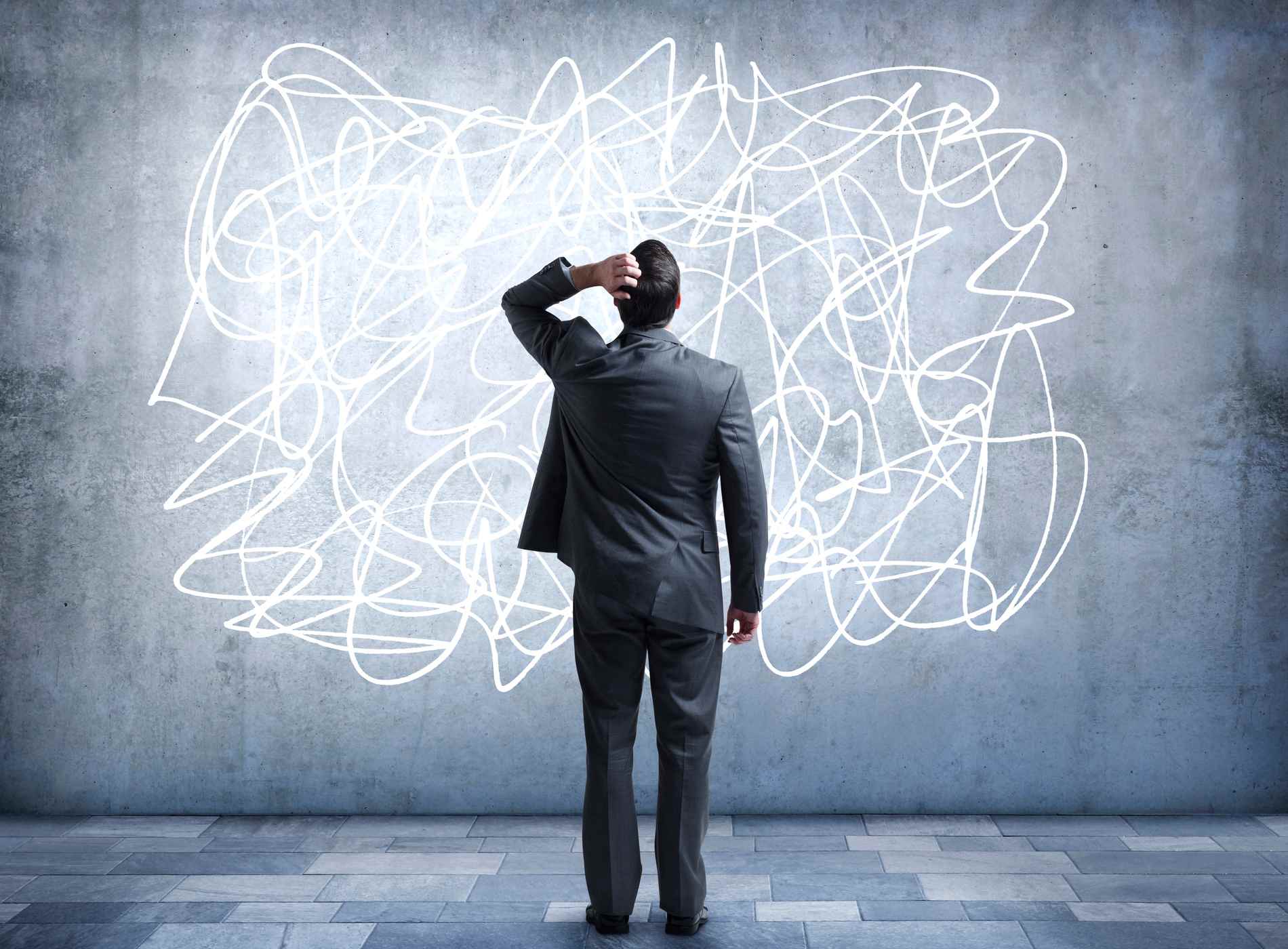 A confused businessman stares at a scribble design that is painted on a wall. It represents the confusion and stress that some people feel in everyday life. He is looking up while standing with his back to the camera.
