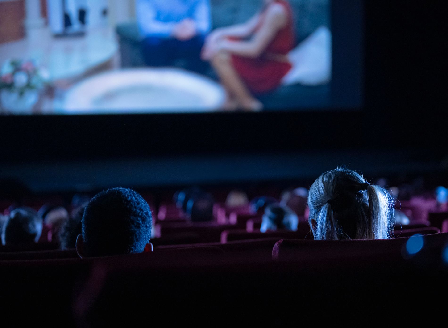 Shot of a movie theater reveals a diverse audience engrossed in a film, with a woman in a red dress on the screen.