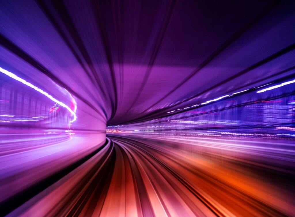 Long-exposure view of a high-speed monorail curving through a neon-lit tunnel at night, with vibrant purple and orange light trails conveying motion, speed and futuristic urban transit.