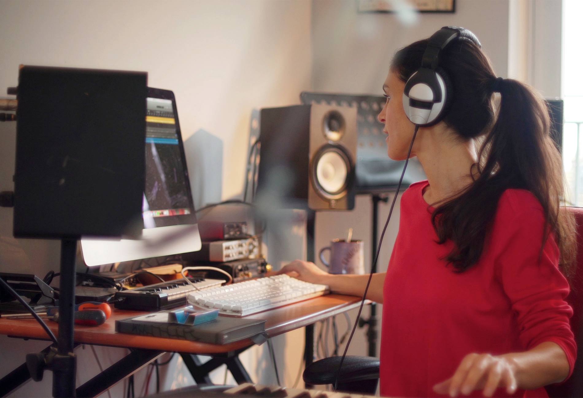 A good looking Hispanic woman is composing music in her private home studio. She is using a computer & music keyboard to compose and is surrounded by musicians’ equipment.