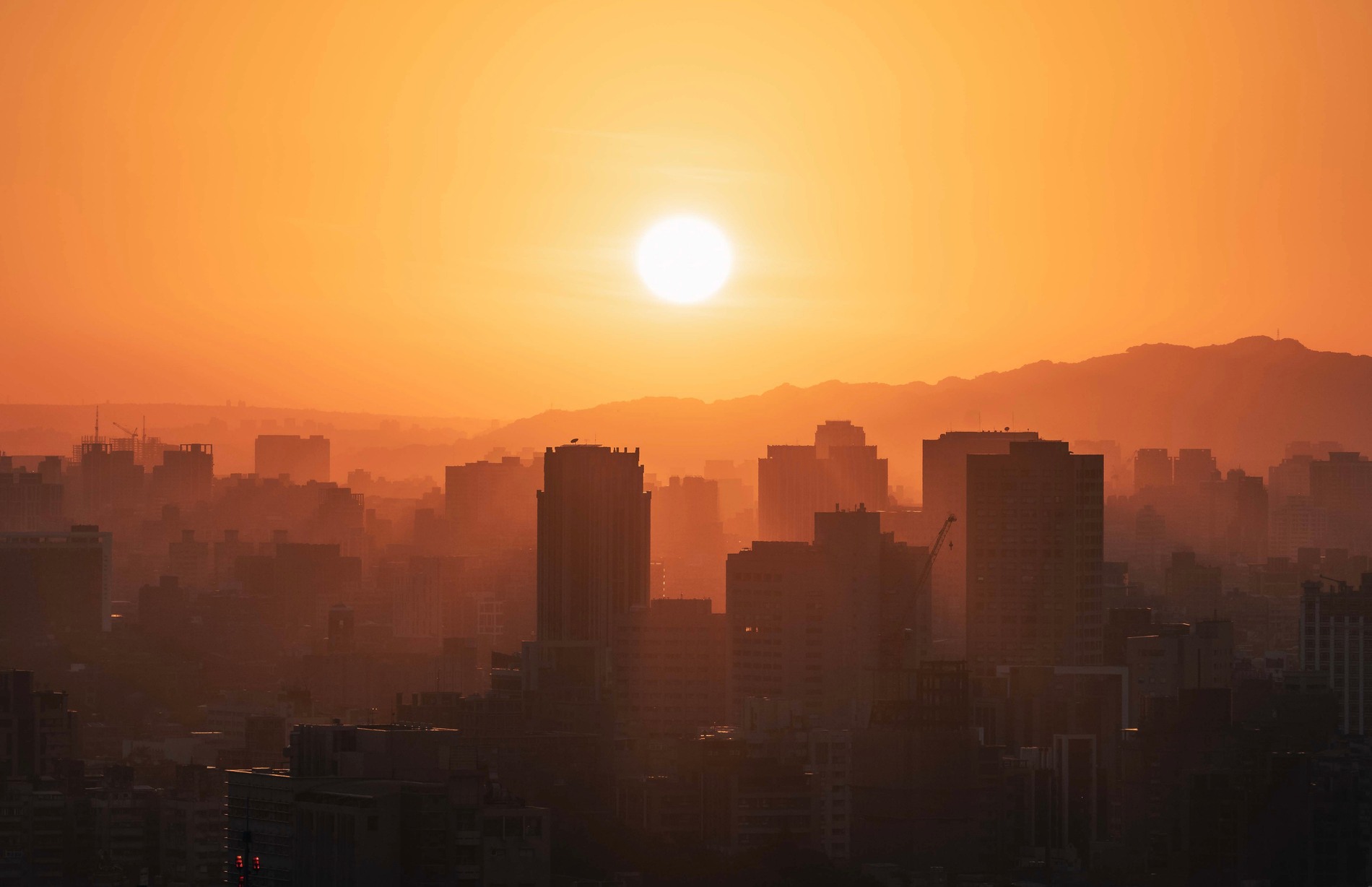 Urban city skyline at sunset covered in heavy smog and air pollution. Silhouetted high-rise buildings under orange sky with visible haze and atmospheric pollution. Concept of climate change, global warming, environmental crisis, heatwave, urban density and air quality issues.